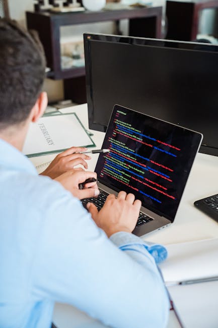 A person working at a desk with multiple monitors and a laptop displaying lines of colorful code. The workspace includes a desktop computer, a notepad, and various office supplies. The background feat