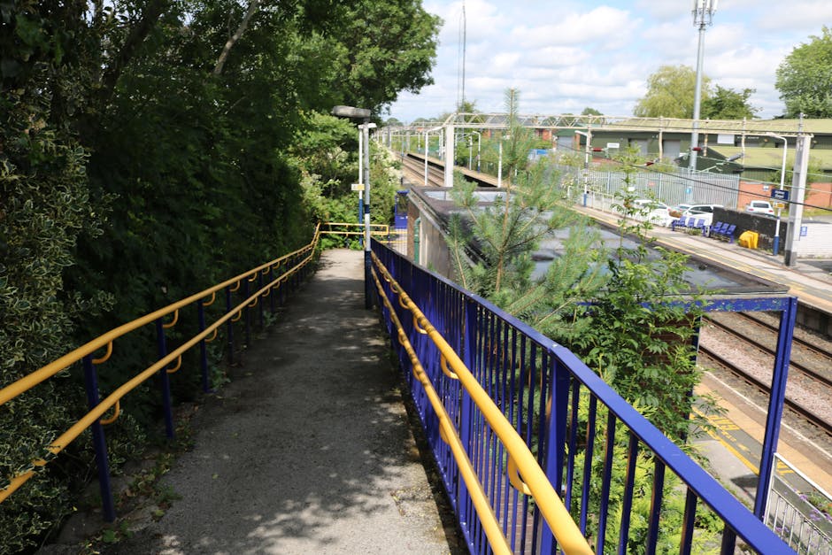 An outdoor pathway running parallel to railway tracks, bordered by dense greenery on the left side with bushes and trees, providing shade and a natural backdrop. The pathway is paved with asphalt and